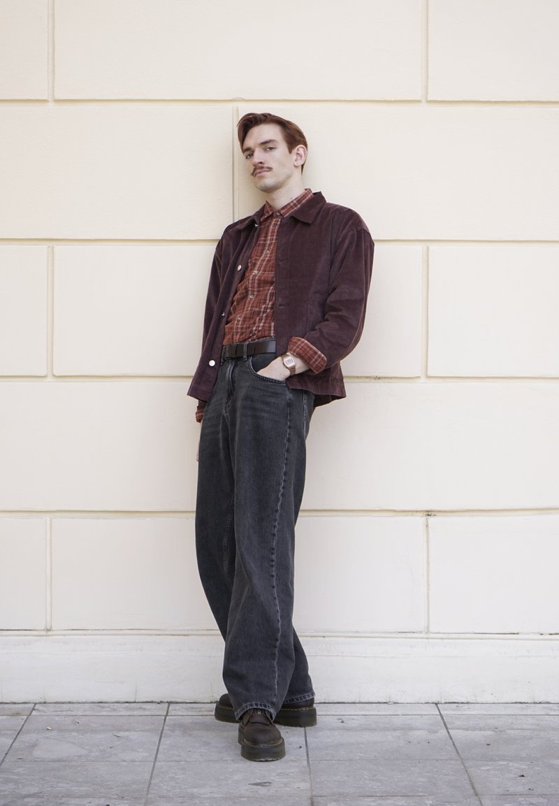 Joven con bigote que lleva una chaqueta granate, camisa de cuadros roja, vaqueros negros sueltos y zapatos negros, apoyado contra una pared de azulejos crema.