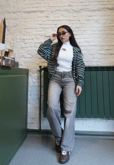 Mujer ajustándose las gafas de sol, lleva chaqueta a rayas, camiseta blanca, vaqueros grises y zapatillas, de pie en el interior contra una pared de ladrillo blanca y un radiador verde.