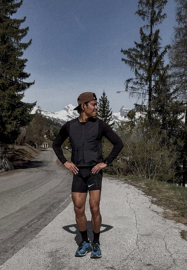 Homme en tenue de sport et casquette debout sur une route de montagne, mains sur les hanches, sommets enneigés et arbres persistants sur fond de ciel clair.
