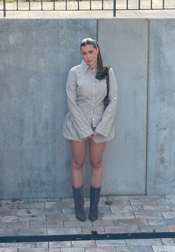 Ženska z dolgimi temnimi lasmi v kare vzorčasti obleki z gumbi in sivimi škornji stoji ob betonskem zidu na tlakovanem površju in gleda naravnost.