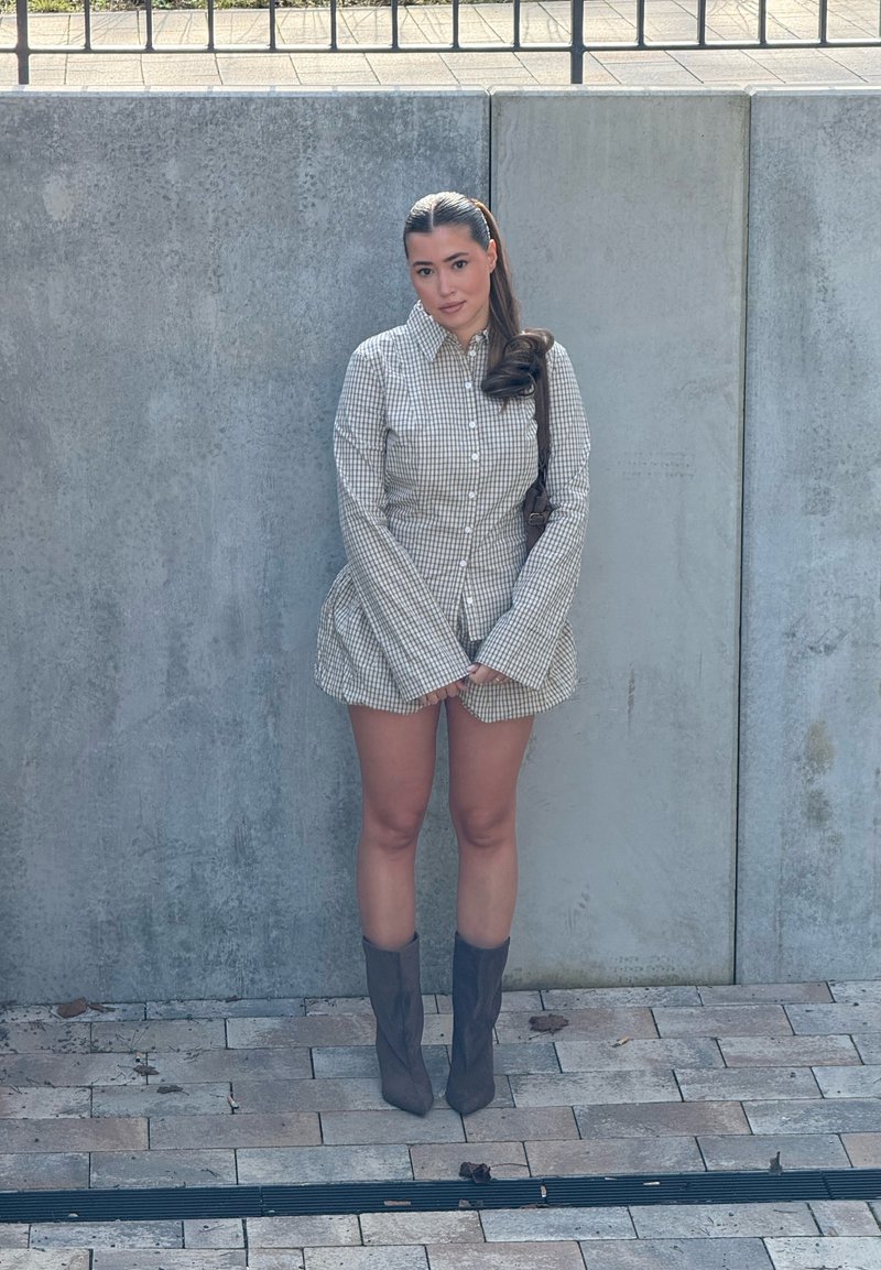 Femme aux longs cheveux noirs portant une robe à carreaux boutonnée et des bottes grises, debout contre un mur en béton sur une surface pavée, regardant devant elle.