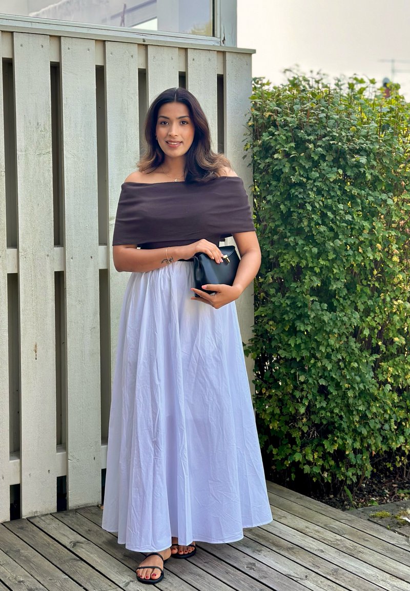 Off-the-shoulder σκούρο καφέ τοπ συνδυασμένο με μια ρέουσα λευκή μίντι φούστα. Μαύρη τσάντα χειρός και μαύρες σαγιονάρες ολοκληρώνουν το σύνολο. Πράσινη φυλλωσιά στο φόντο.