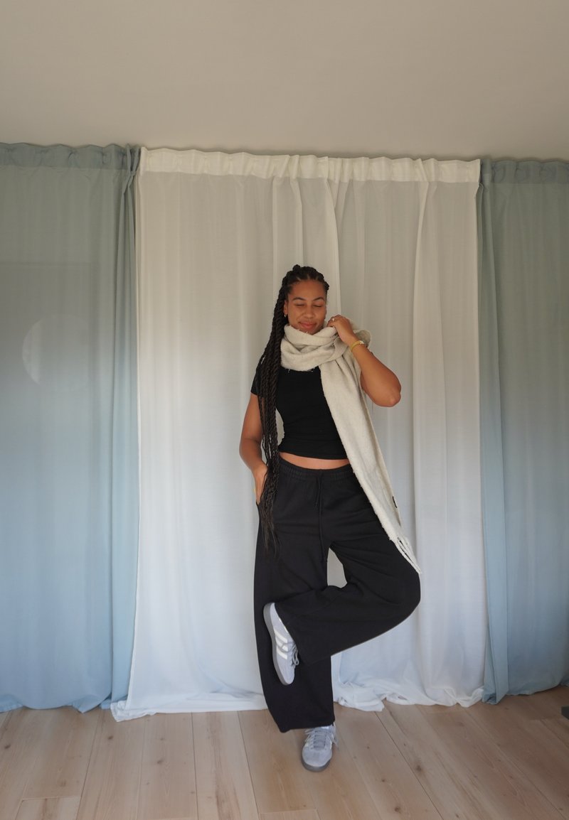 Calças de moletão oversized pretas com um top cropped preto, cachecol cinza e ténis cinzas num chão de madeira contra cortinas claras.
