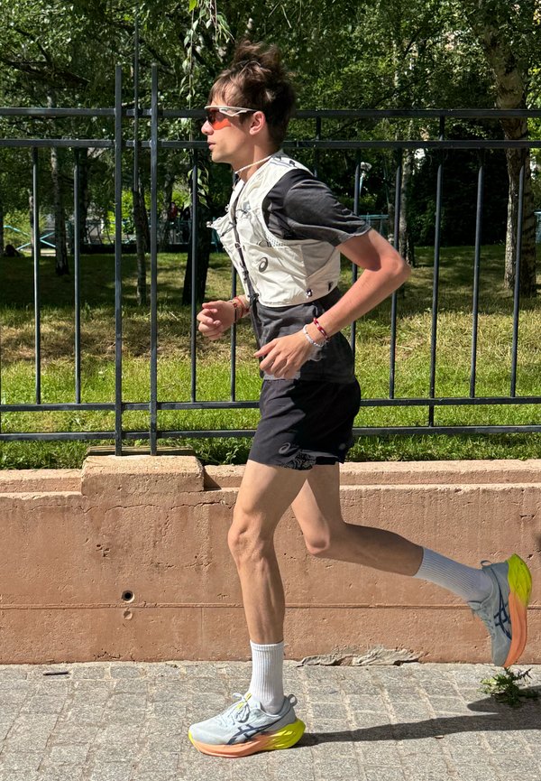Jeune homme courant en plein air portant des lunettes de soleil, un gilet d'hydratation, un short noir, des chaussettes blanches et des chaussures de course grises avec des semelles jaunes vives.