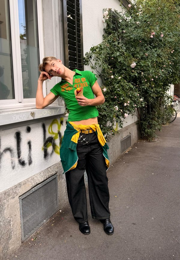 Junge Person in grünem Hemd und schwarzen Hosen lehnt an einer mit Graffiti bedeckten Wand und hält ein Eishörnchen neben einem Kletterrosenstrauch auf dem Gehweg.