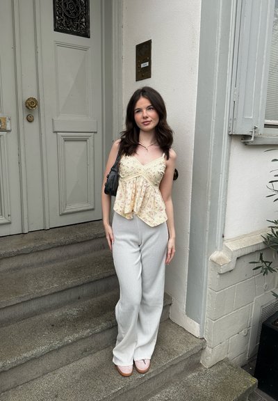 Top peplum amarillo floral con tirantes finos, combinado con pantalones de pierna ancha gris claro y zapatos rosas. De pie en unos escalones de piedra junto a una puerta.