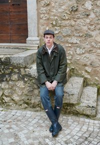 Zelená vodotěsná bunda, bílá košile, modré džíny a černé gumové boty. Osoba sedící na kamenných schodech u texturované kamenné zdi.