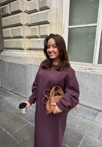 Lang, oversized, paarse sweatshirtjurk gemaakt van zachte stof, gecombineerd met een tan lederen tas en een zwart wegwerp koffiekopje.