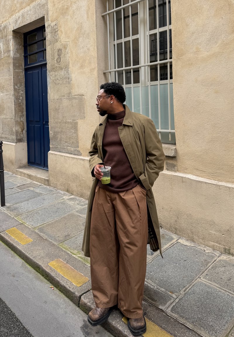 Homme en col roulé marron, trench-coat beige et pantalon large tenant une boisson verte, debout sur un trottoir en ville près d’un mur beige et d’une porte bleue.