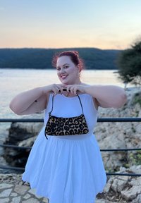Taschen mit Leopardenmuster in geschwungener Form und schwarzem Riemen, gehalten von einer Person in einem weißen Kleid, vor einer Wasserlandschaft.
