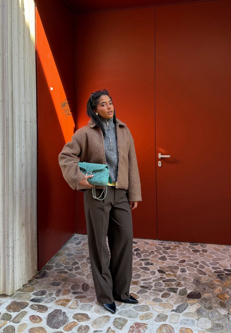 Femme debout sur un pavé en pierre, portant une veste teddy marron, un pull gris, un pantalon foncé, des chaussures noires pointues, et tenant un sac matelassé bleu sarcelle.