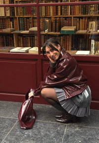 Bordo odinė striukė, pilkas plisė sijonas, juodi aukštakulniai iki šlaunų ir blizgi bordo spalvos rankinė, nufotografuoti prie spalvingų knygų fono.