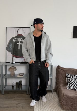 Hombre con gorra negra, sudadera gris con cremallera, camiseta negra y pantalones negros holgados está de pie en una habitación con un cuadro enmarcado de una chaqueta, una estantería y un sofá con un cojín de cebra.