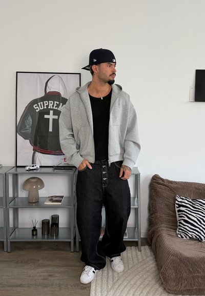 Hombre con gorra negra, sudadera gris con cremallera, camiseta negra y pantalones negros holgados está de pie en una habitación con un cuadro enmarcado de una chaqueta, una estantería y un sofá con un cojín de cebra.
