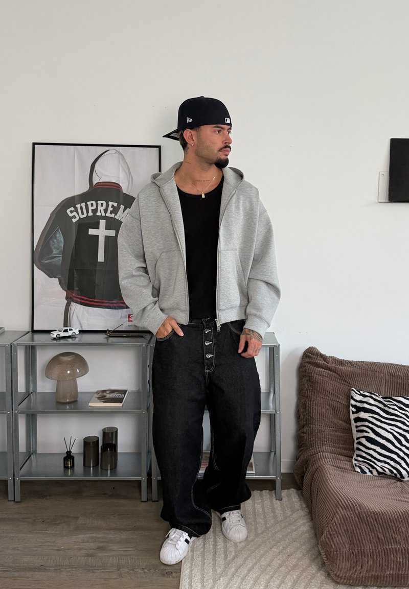 Homme avec casquette noire, sweat à capuche gris zippé, t-shirt noir et pantalon noir ample, debout dans une pièce avec une œuvre encadrée de veste, étagère et canapé avec coussin zèbre.