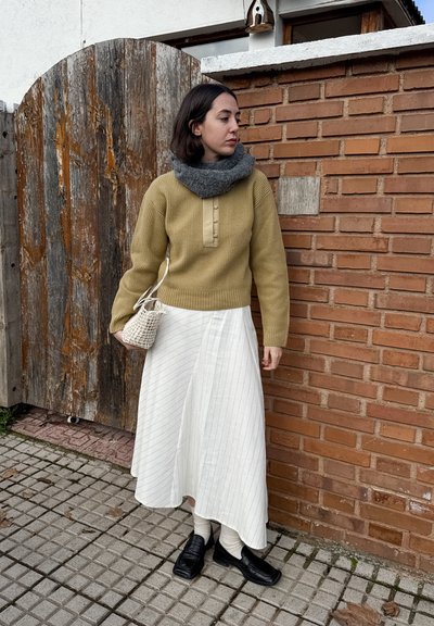 Mujer con suéter verde oliva, falda blanca, bufanda gris y zapatos negros, sosteniendo un bolso blanco tejido, de pie junto a una pared de ladrillos y una puerta de madera al aire libre.