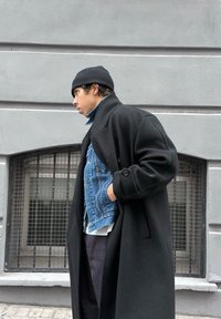 Νεαρός άνδρας με μαύρο σκουφάκι, μακρύ μαύρο παλτό και τζιν μπουφάν, στέκεται πλάγια σε γκρι τοίχο με πανοπλίες στα παράθυρα.