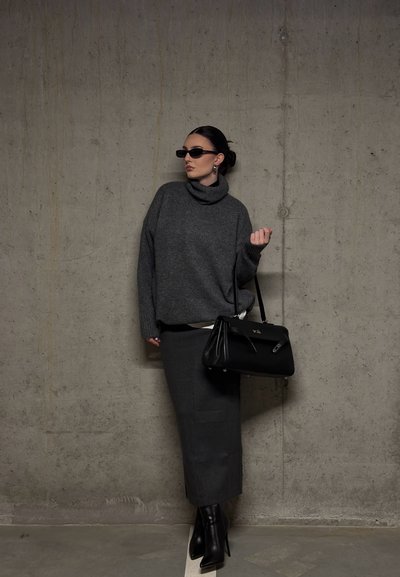 Mujer con suéter de cuello alto gris oscuro, falda larga a juego, botas negras con tacón, gafas de sol y sosteniendo un bolso negro junto a una pared de concreto.