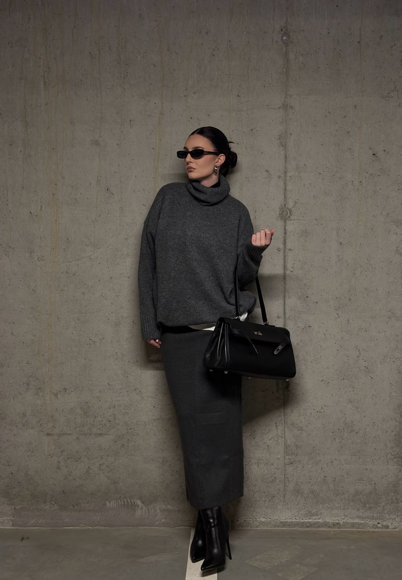 Donna con maglione a collo alto grigio scuro, gonna lunga coordinata, stivali neri con tacco, occhiali da sole e una borsa nera, appoggiata a un muro di cemento.