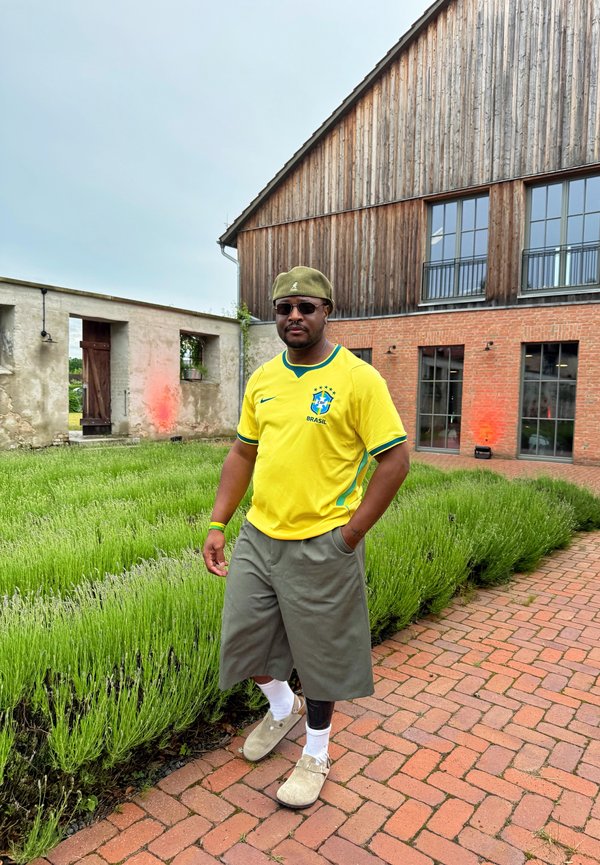 Mand iført en gul Brasiliens fodboldtrøje, grå shorts, grøn hat og solbriller går på en brostenssti ved siden af grønne planter og rustikke bygninger.