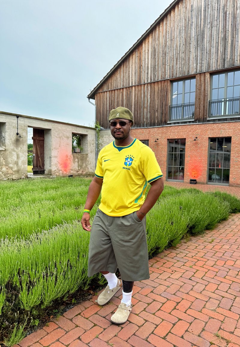 Homme portant un maillot de football jaune du Brésil, un short gris, un chapeau vert et des lunettes de soleil, marchant sur un chemin en briques à côté de plantes vertes et de bâtiments rustiques.