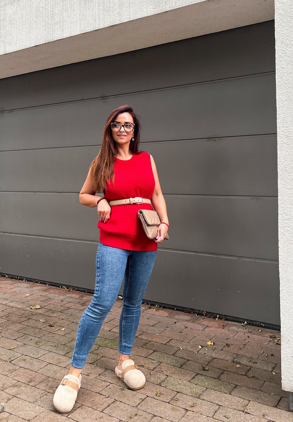 Röd stickad ärmlös topp, slimmade blå jeans, beige bälte, brun clutchväska och beige tofflor, mot en grå vägg och tegelpavement.