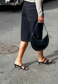 Μαύρα τζιν σορτς με άνετη εφαρμογή, συνδυασμένα με μαύρες σαγιονάρες με τακούνι και μια καμπύλη μαύρη τσάντα, καθώς περπατούν σε μια γκρίζα επιφάνεια.