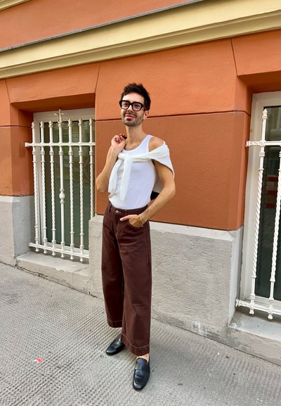 Top sin mangas blanco con suéter de punto blanco drapeado sobre los hombros; pantalones de pierna ancha marrones; mocasines negros; de pie contra una pared naranja.