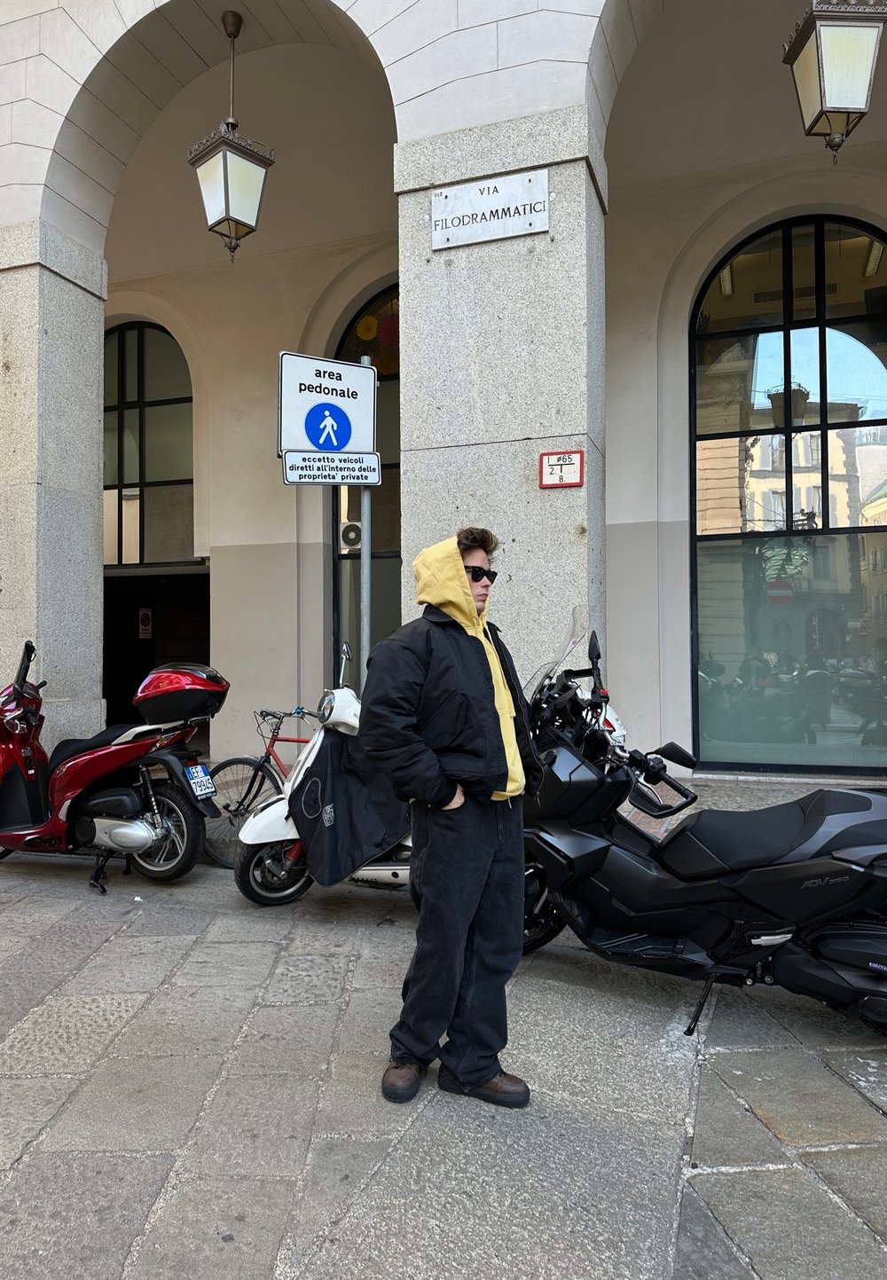 Posameznik v črni puhovki čez rumeno kapuco, temnimi hlačami in temnimi čevlji, stoji blizu parkiranih skuterjev pod lokom.