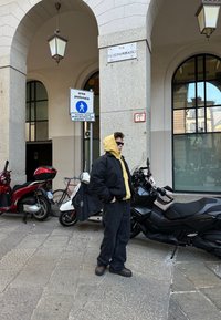 Posameznik v črni puhovki čez rumeno kapuco, temnimi hlačami in temnimi čevlji, stoji blizu parkiranih skuterjev pod lokom.