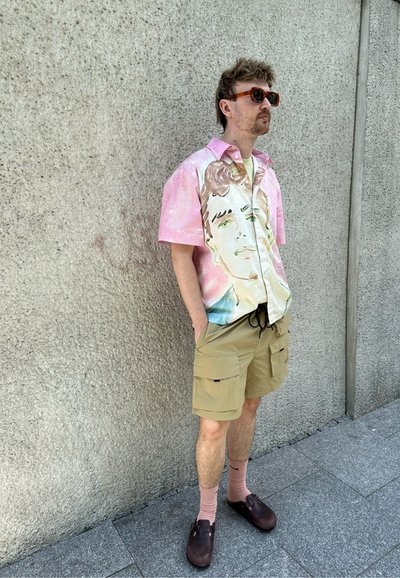 Camisa de manga corta, rosa y beige, con un diseño gráfico, combinada con pantalones cortos de carga caqui, calcetines rosas y zuecos marrones.