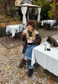 En kvinde iført en brun lammeskindsfrakke og pels hat sidder ved et bord og holder en kaffekop, med en tallerken med kager foran sig.