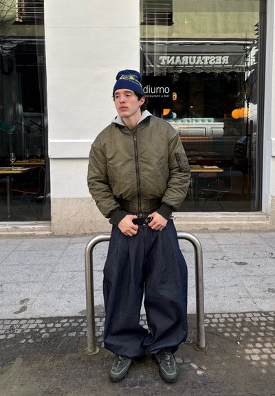 Joven con chaqueta bomber verde, gorro azul y pantalones anchos oscuros, de pie en la acera frente a un restaurante con ventana de cristal.