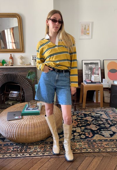 Camisa de manga larga a rayas amarillas y verdes, pantalones cortos de mezclilla azules, botas de rodilla color beige, ubicados en una habitación decorada con una mesa redonda de mimbre.
