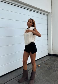 Maglione bianco lavorato a maglia con coste, pantaloni corti neri e stivali grigi fino al ginocchio. Posizione in piedi davanti a uno sfondo di porta del garage bianca.
