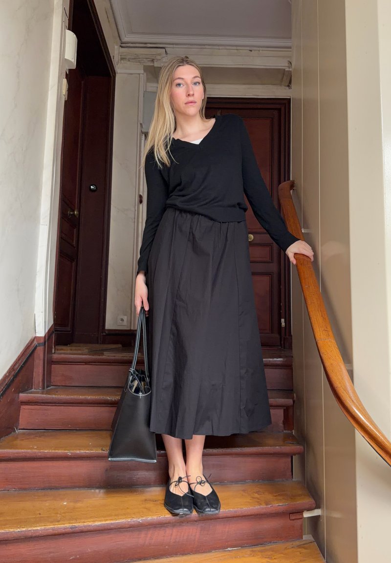 Jeune femme portant un haut noir à manches longues, une jupe noire et des ballerines noires, debout sur un escalier en bois, tenant un sac à main noir et touchant la rampe.