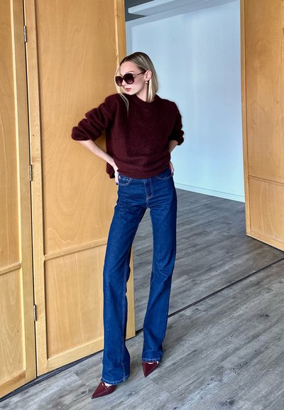 Jersey esponjoso color burdeos, jeans de tiro alto azules y tacones burgundy en punta; de pie frente a puertas de madera en un entorno minimalista.