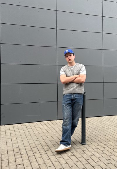 Joven con chaleco de punto gris, gorra azul de Los Ángeles, vaqueros y zapatillas blancas apoyado contra un poste negro frente a una pared gris con azulejos.