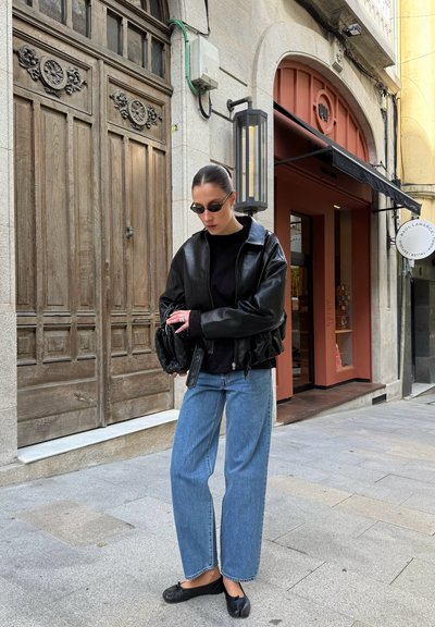 Mujer que lleva una chaqueta de cuero negra, top negro, gafas de sol, vaqueros azules y zapatos negros, de pie en una calle de la ciudad cerca de una puerta de madera y la entrada de una tienda.