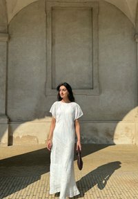 Weißes Maxikleid mit kurzen, weiten Ärmeln, das über ein dezentes texturiertes Muster verfügt. Getragen mit einer dunklen, strukturierten Clutch in einem sonnendurchfluteten Ambiente.
