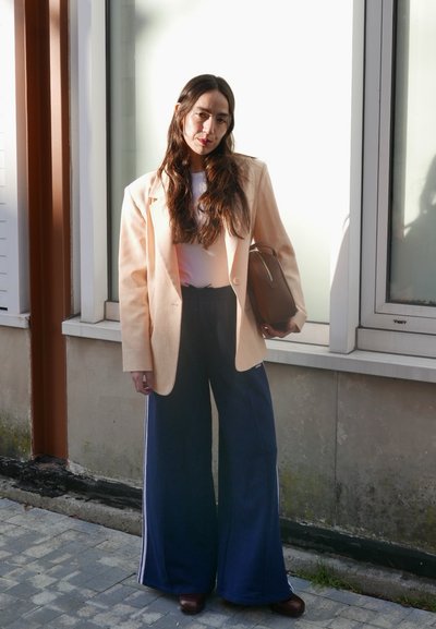 Blazer melocotón sobre una blusa blanca, combinado con pantalones de pierna ancha en azul marino. Bolso marrón, zapatos marrones y cabello largo y ondulado. Entorno luminoso al aire libre.
