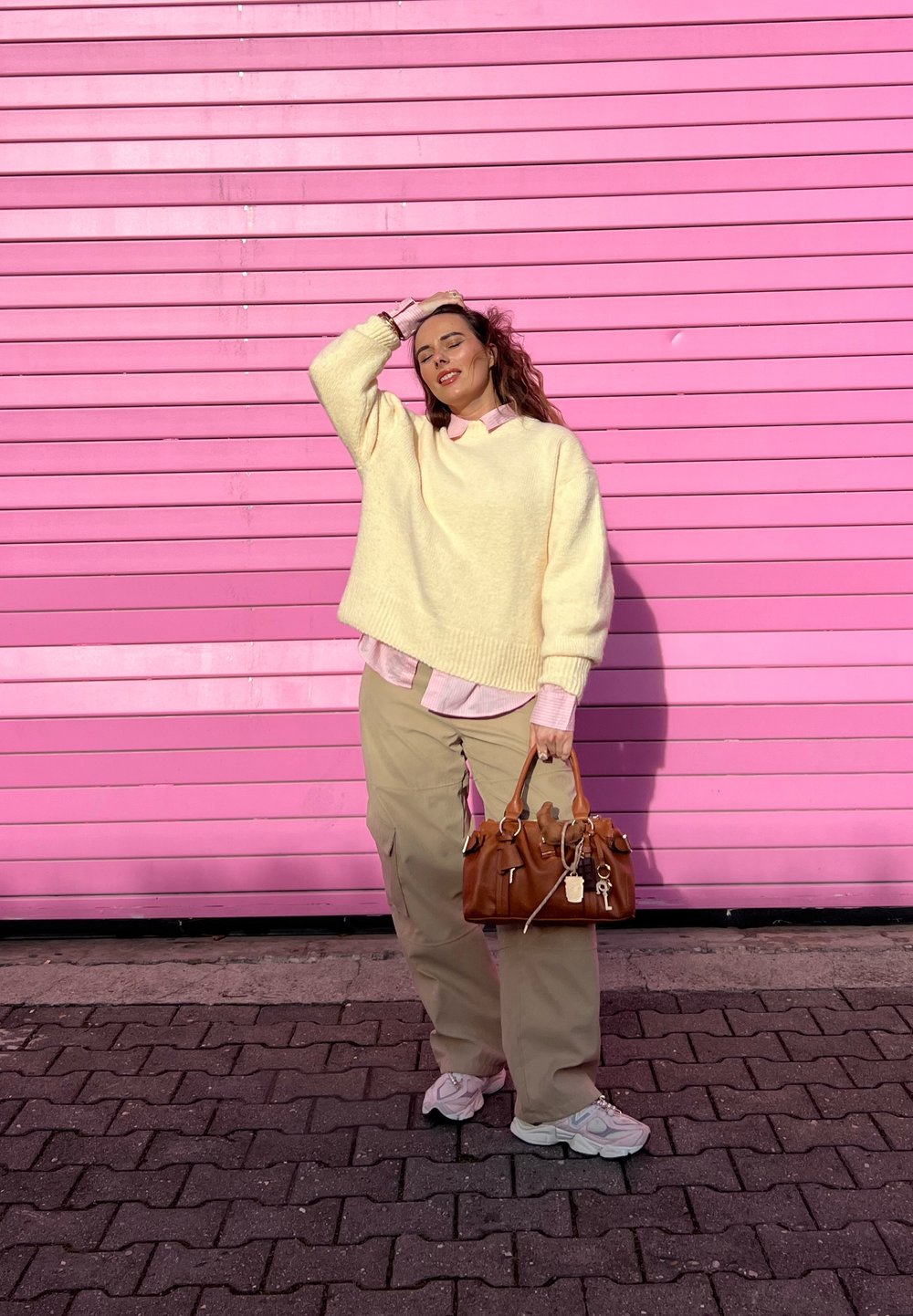 Pull texturé crème porté sur une chemise légère rose pâle à col, associé à un pantalon cargo kaki et un sac à main marron avec des accents métalliques.