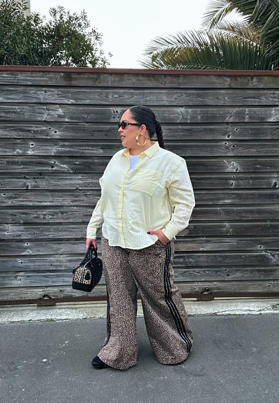 Mujer con cabello trenzado, lleva gafas de sol, pendientes de aro, camiseta amarilla, pantalones anchos con estampado de leopardo, sostiene un bolso negro y con estampado de leopardo, está de pie al aire libre.