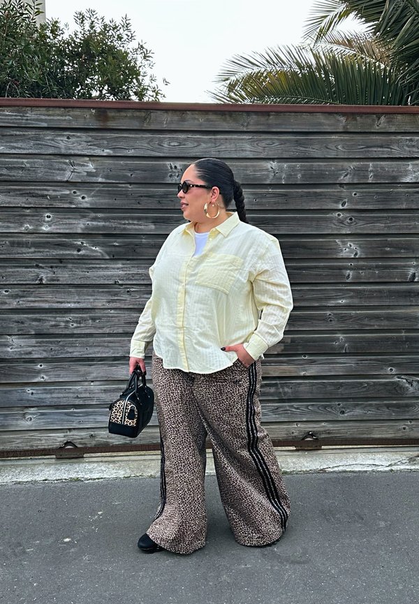 Femeie cu părul împletit purtând ochelari de soare, cercei cerc, cămașă galbenă, pantaloni largi cu imprimeu de leopard, ținând o geantă neagră și cu imprimeu de leopard, stând în aer liber.