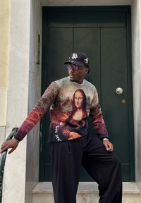 Pull avec une impression de la Joconde aux couleurs vives, à manches longues. Associé à un pantalon noir ample et une casquette.