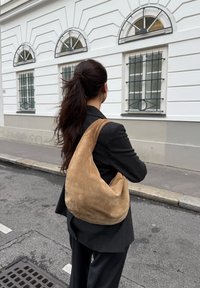 Peščena suedelna torba za rame z mehkimi teksturami, hobo obliko in enim širokim trakom, postavljena na nevtralen ozadje.