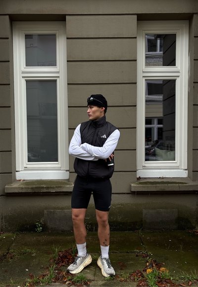 Persona con chaleco negro, mangas largas blancas, pantalones cortos negros y zapatillas, de pie con los brazos cruzados entre dos ventanas en una pared gris.