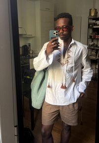 White long-sleeve shirt with brown embroidery and tassels, paired with brown shorts. Holding a turquoise bag in a bright interior.