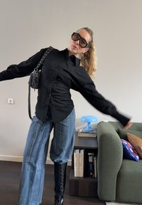 Femme portant une chemise noire surdimensionnée, un jean bleu, de grandes bottes noires et de grandes lunettes de soleil, posant avec les bras écartés dans un salon.