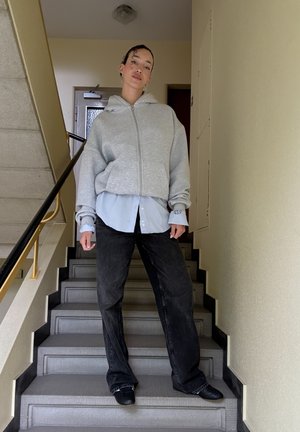 Sudadera gris con cremallera sobre una camisa de botones azul claro, combinada con pantalones vaqueros de pierna ancha oscuros y zapatos negros sin cordones, de pie en una escalera.